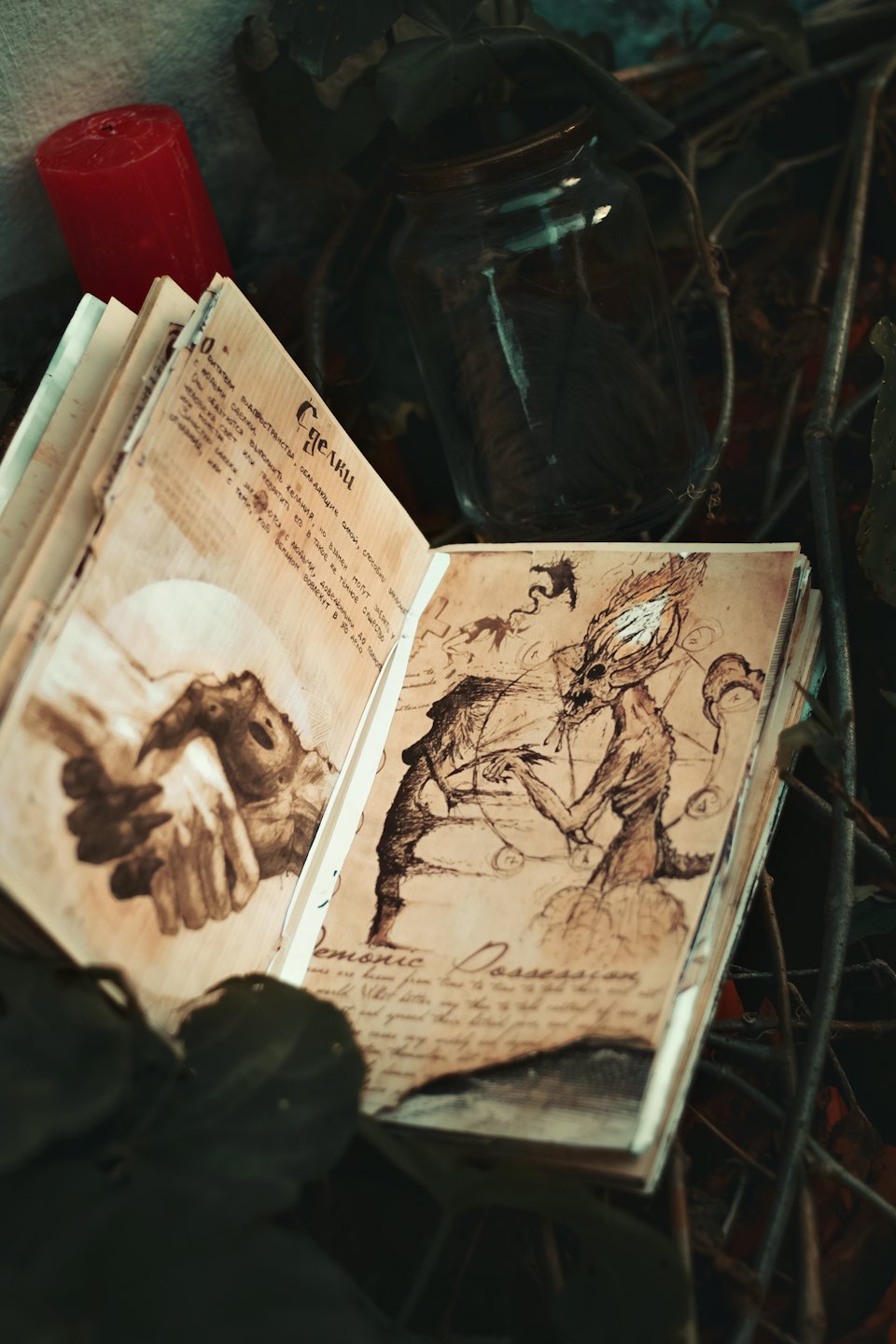 Livro antigo aberto sobre galhos secos, com desenhos a tinta de uma criatura demoníaca ao lado de uma vela