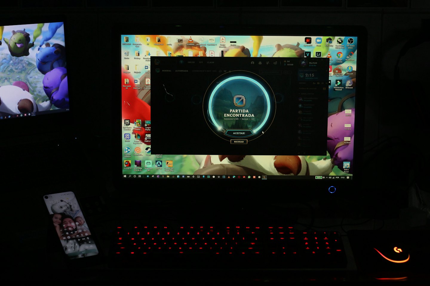 Setup de jogo com dois monitores, teclado e celular sobre a mesa.